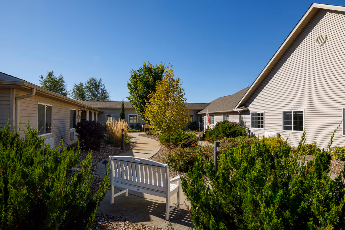 Sunlit courtyard with a white bench, winding concrete path, shrubs and trees between single-story beige buildings under a clear blue sky.