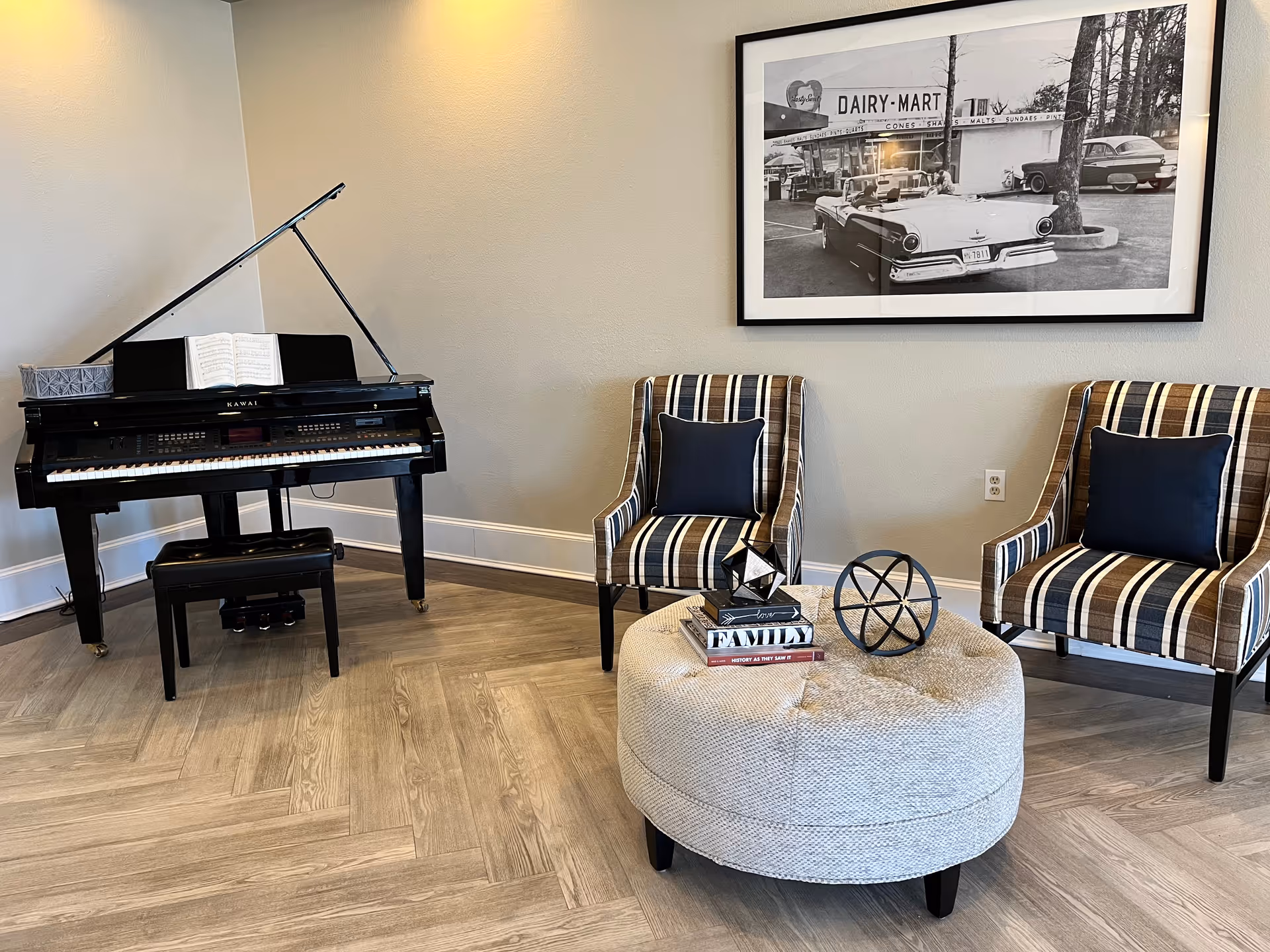 A cozy interior corner featuring a black grand piano with an open sheet of music, two striped armchairs with dark cushions, and a round beige ottoman with decorative items and books on top. A large black and white framed photograph of vintage cars and a Dairy-Mart sign hangs on the wall behind the chairs.