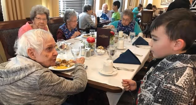 A group of elderly people sitting around a dining table in a senior living facility, eating and conversing. A young boy stands nearby, engaging with one of the elderly individuals. The table is set with glasses, cups, silverware, and a menu in the center.