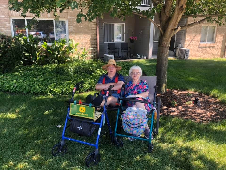Two elderly individuals sitting outdoors on a bench under a tree in a grassy area. Both have walkers in front of them, and behind them is a building with windows and a small patio area with chairs and potted plants.