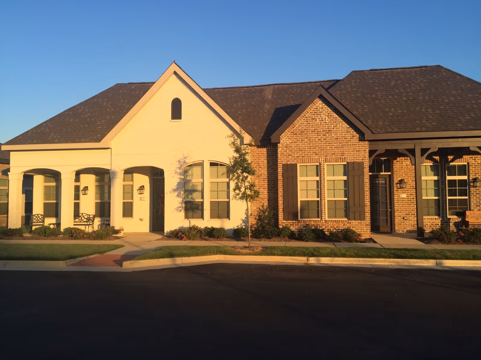 Exterior view of a single-story building with a combination of white and red brick walls, multiple windows with shutters, a dark shingled roof, and a small tree and landscaping in front. The building has a covered porch area with benches and is illuminated by warm sunlight under a clear blue sky.
