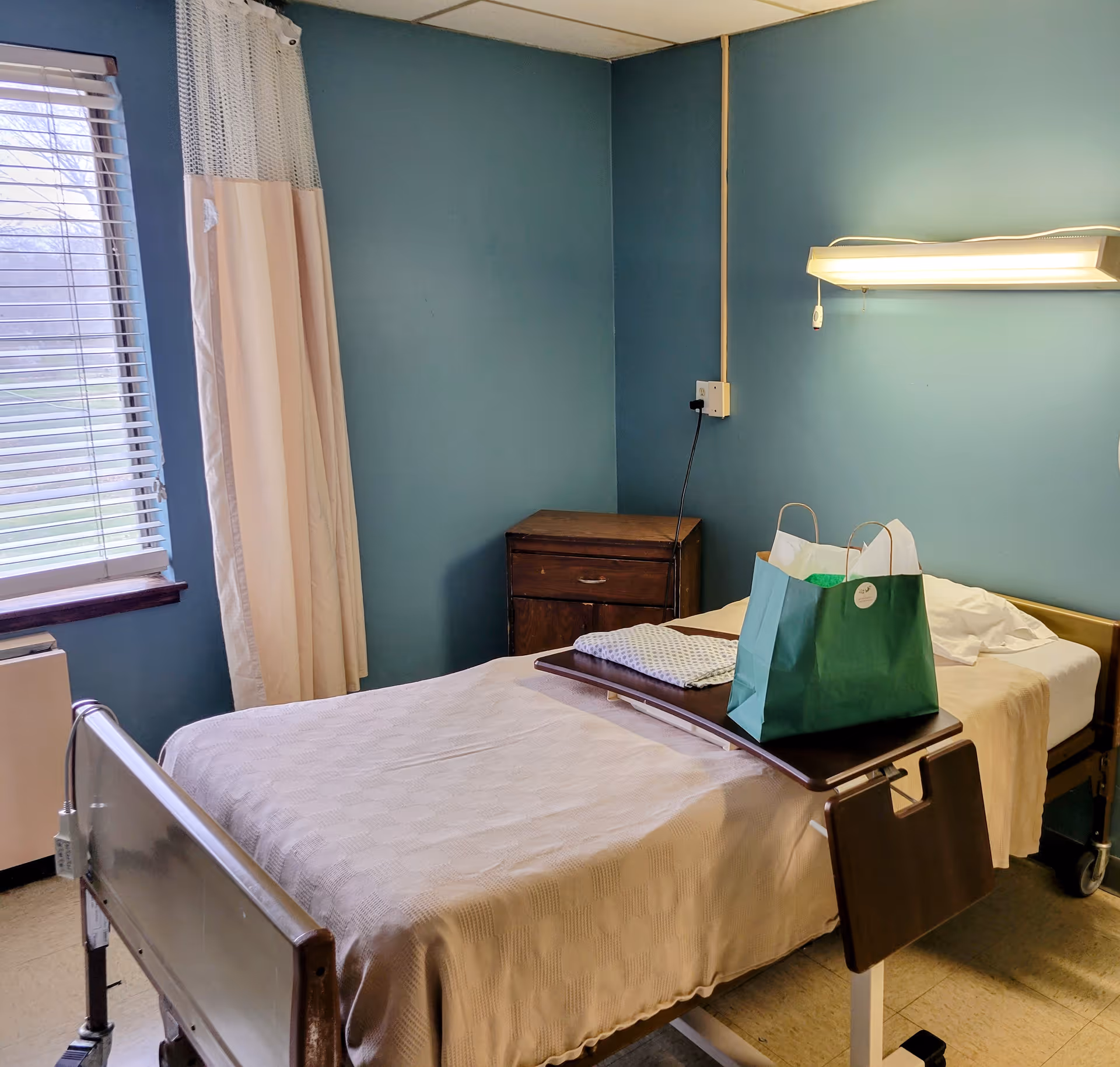 A small healthcare facility bedroom with a single hospital bed covered with a beige blanket. A green paper bag and folded blanket are placed on a wooden overbed table. The room has teal walls, a window with blinds and curtains, a wooden nightstand, and a wall-mounted light fixture above the bed.