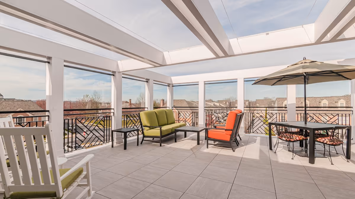 A spacious outdoor patio area with a white pergola overhead, featuring a green cushioned sofa, an orange cushioned chair, a small black coffee table, a black dining table with four chairs, and a large umbrella providing shade. The patio overlooks rooftops and trees under a partly cloudy sky.