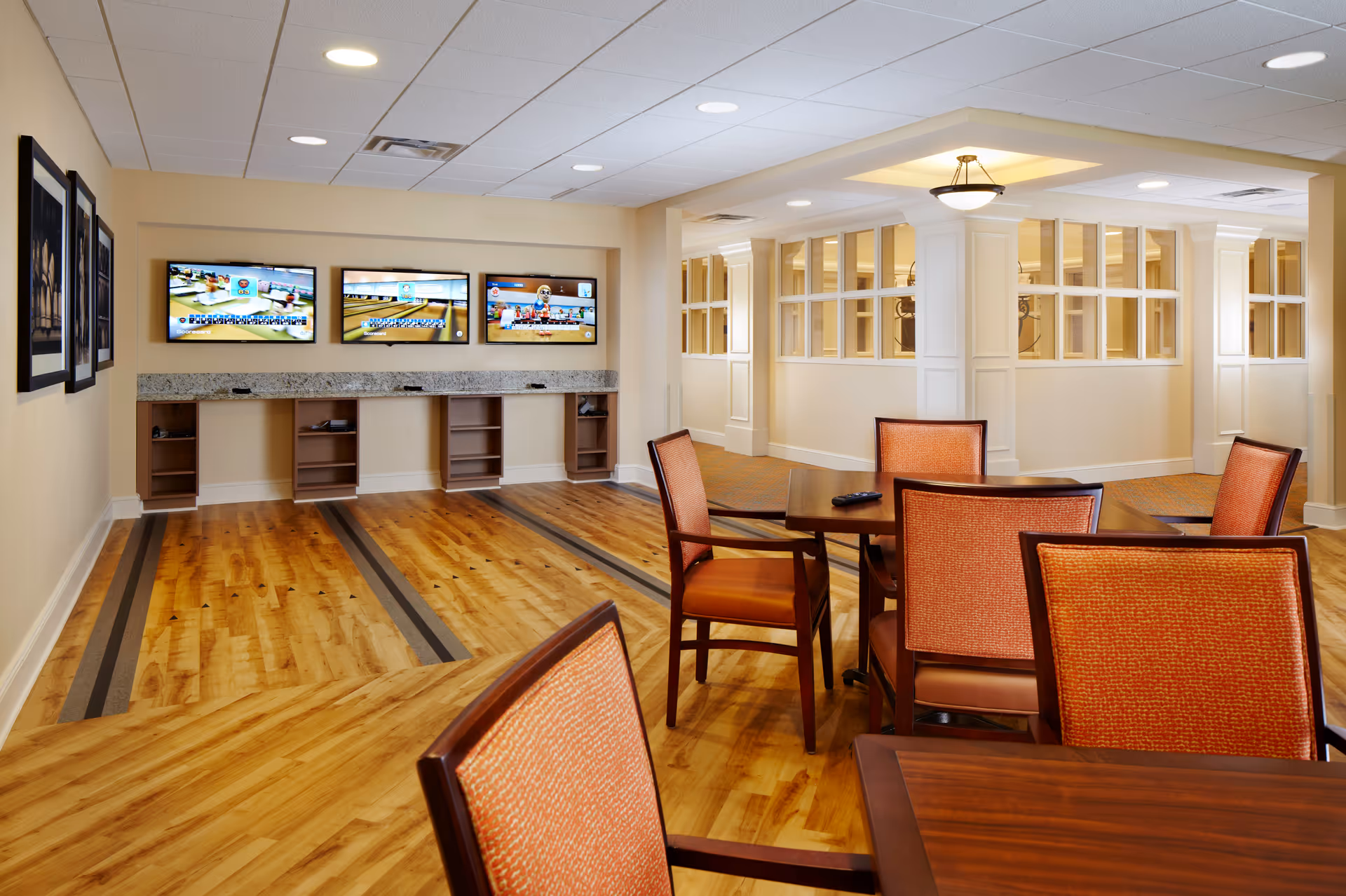 Interior view of a senior living facility game room with a wooden floor designed for bowling. The room features three wall-mounted flat-screen TVs above a granite countertop with open shelving underneath. There are several orange-upholstered chairs around wooden tables in the foreground. The walls are light-colored with framed pictures and windows with white trim in the background.