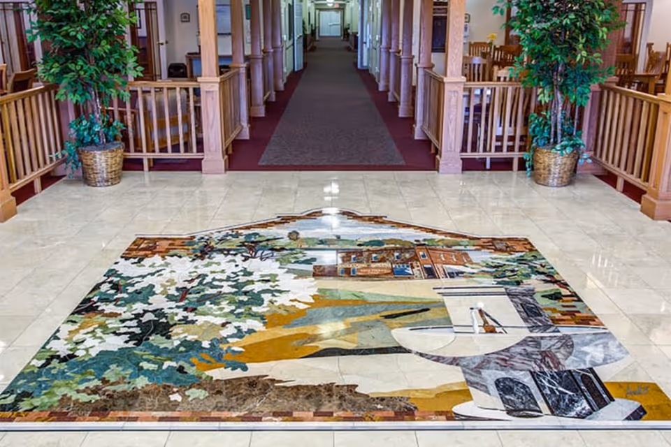 Interior view of a senior living facility hallway with a decorative floor mosaic depicting a scenic landscape. The hallway is lined with wooden railings and potted plants on either side, leading to a carpeted corridor with doors and seating areas visible in the background.