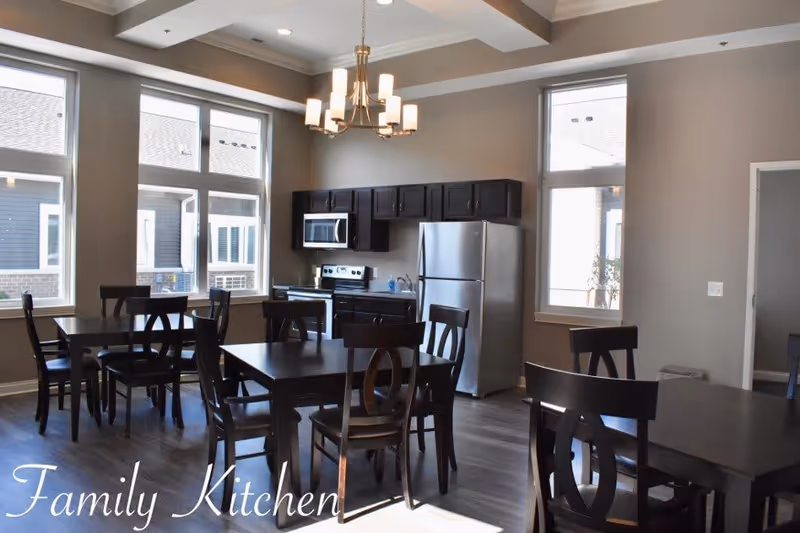 A bright kitchen and dining area with multiple dark wooden tables and chairs. The kitchen features dark cabinetry, a stainless steel refrigerator, microwave, and stove. Large windows allow natural light to fill the room, and a modern chandelier hangs from the ceiling.