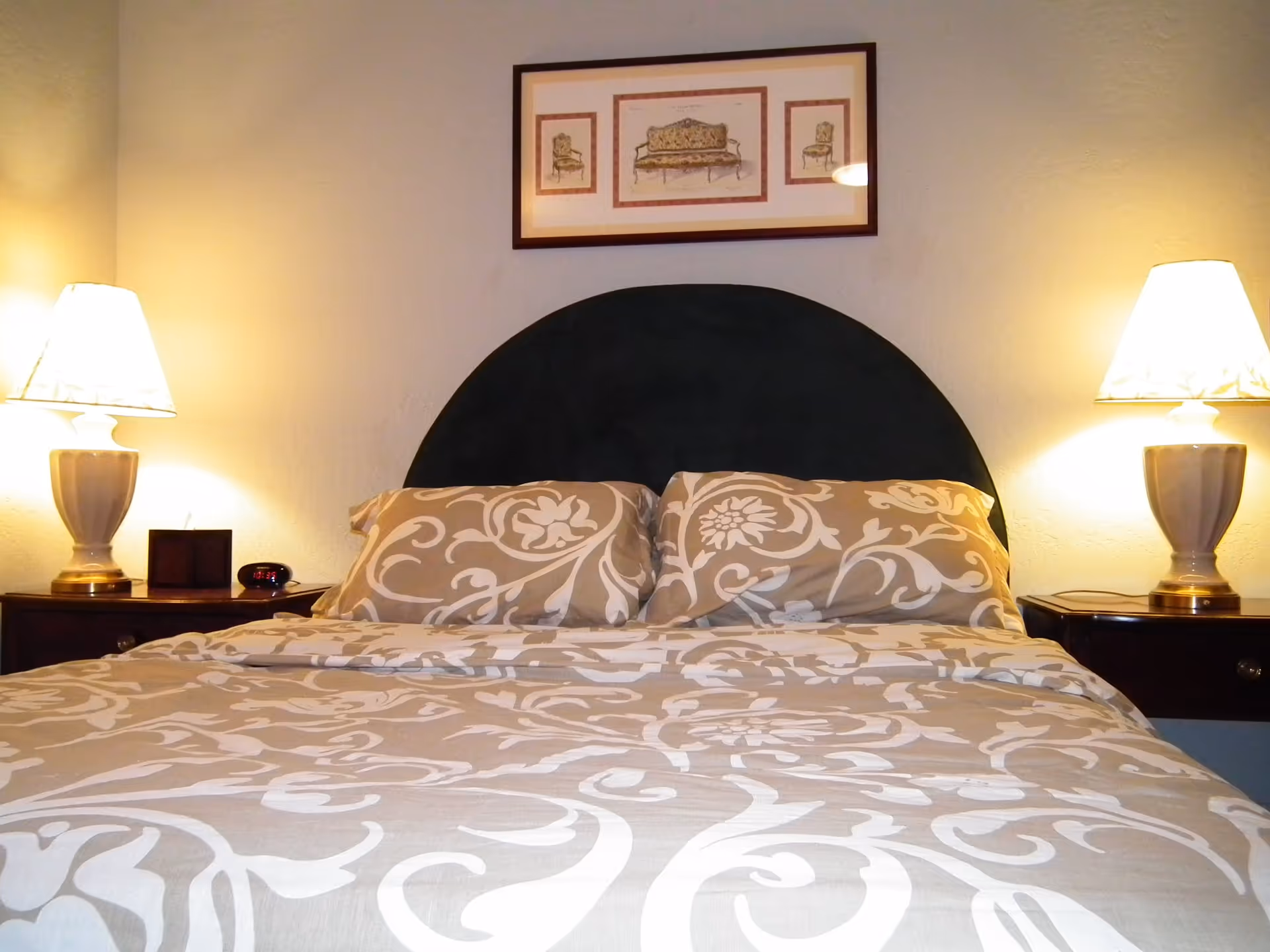 Well-lit bedroom with a patterned bed, a dark rounded headboard, two bedside lamps and a framed picture above.
