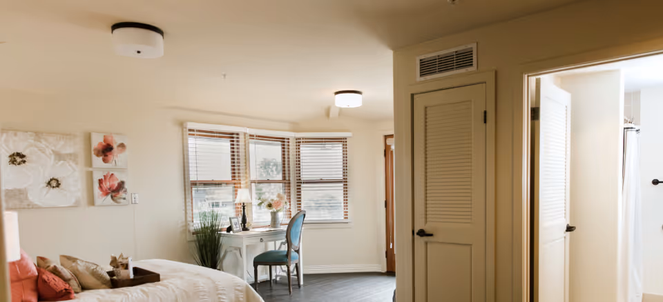 A cozy bedroom with a bed on the left side adorned with pillows and a tray, floral wall art above the bed, a white desk with a blue chair in front of a bay window with blinds, and a door leading to a bathroom on the right side.