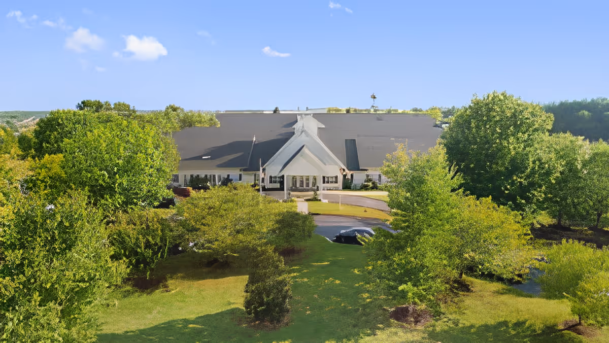 Front exterior of a large assisted living building set behind trees and lawn with a circular driveway and a parked car.