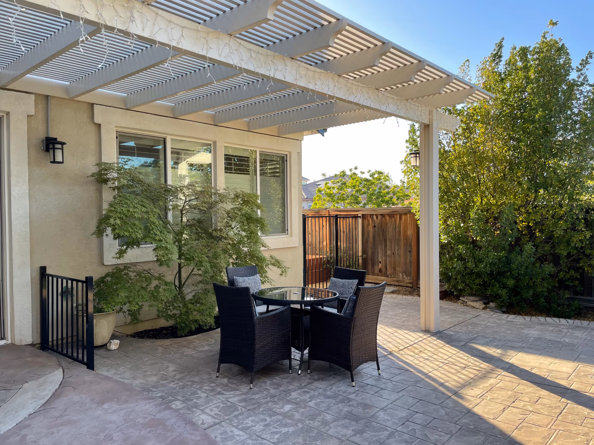 Outdoor patio area at Deer Ridge Country Villa featuring a glass-top round table with four black wicker chairs with cushions. The patio is covered by a white pergola with string lights hanging from it. There are green bushes and trees along the wooden fence surrounding the patio.