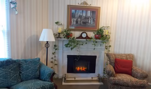 A cozy living room area featuring a white fireplace with a small electric fire, decorated with green plants and a clock on the mantel. Above the fireplace hangs a framed picture. To the left is a blue patterned sofa, and to the right is a patterned armchair with a red cushion. A floor lamp stands next to the sofa, and the walls have light-colored vertical striped wallpaper.