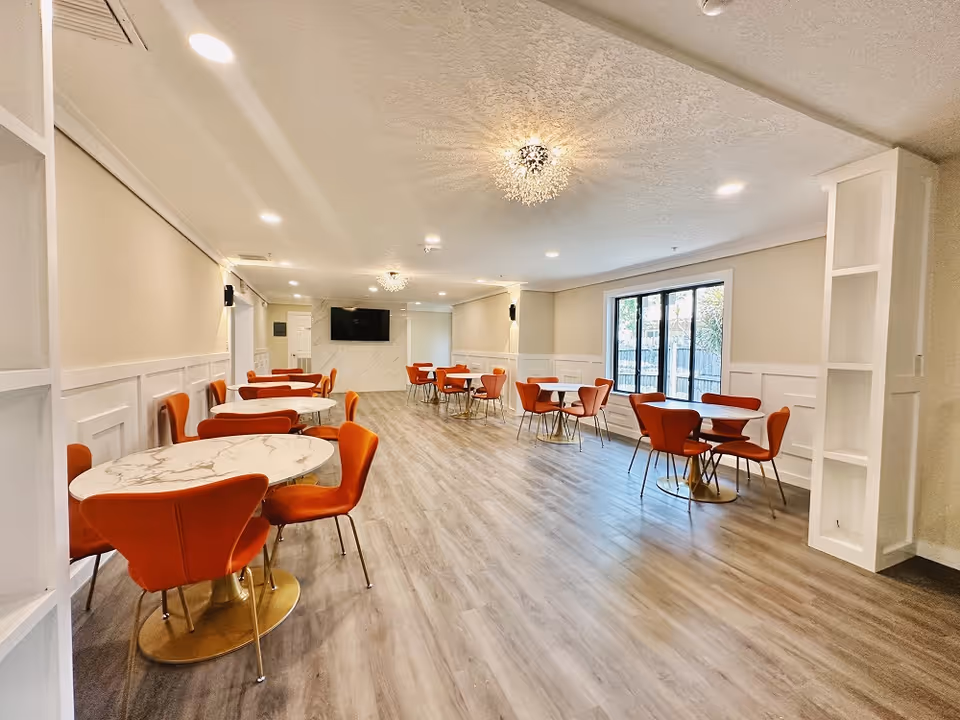 A bright and modern dining room with multiple round marble tables surrounded by orange chairs. The room has light wood flooring, white walls with wainscoting, and large windows letting in natural light. There is a flat-screen TV mounted on a marble accent wall at the far end of the room and decorative ceiling lights.