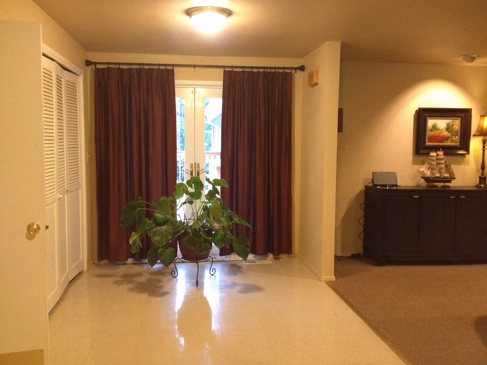 Interior view of a room with a large potted plant on a stand in front of closed maroon curtains covering glass doors. To the right, there is a dark wooden cabinet with a model ship, a framed painting, and a lamp. The floor is tiled in the foreground and carpeted in the background.