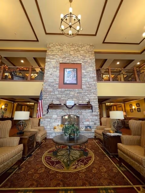A spacious senior living facility common area with a large stone fireplace as the focal point. The room features comfortable armchairs arranged around a round coffee table with a plant centerpiece. There are two table lamps on side tables beside the chairs. The ceiling is high with wooden beams and a chandelier hanging in the center. An American flag is positioned to the left of the fireplace. The upper level has a railing overlooking the area below.
