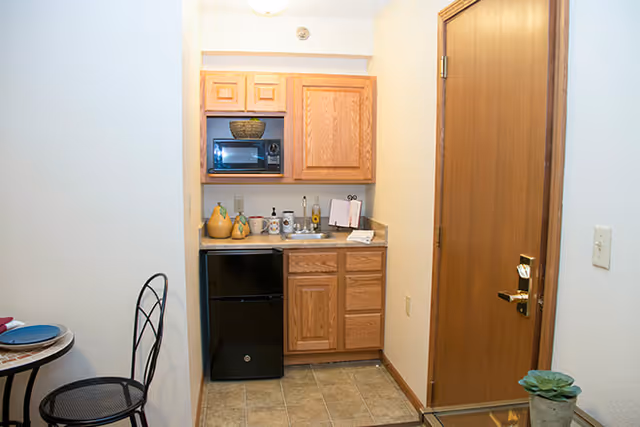 Small kitchenette area with wooden cabinets, a black mini refrigerator, a microwave, and a countertop with various items including a soap dispenser and a small towel. To the right is a wooden door with a gold-colored handle, and to the left is a small round table with a black metal chair.