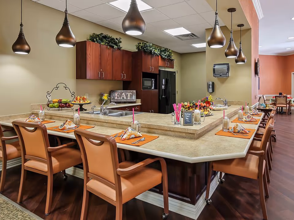A well-lit kitchen and dining area in an assisted living facility featuring a large countertop with multiple place settings. Each setting includes an orange placemat, a folded napkin, a glass with a pink straw, and silverware. The kitchen has wooden cabinets, a black refrigerator, a microwave, and a double sink. Decorative plants and a sign that says 'EAT' are on the countertop. Pendant lights hang from the ceiling above the counter. In the background, there is a dining room with tables and chairs.