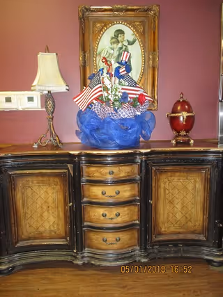 A wooden sideboard with a vintage style, featuring three central drawers and two cabinets on each side. On top of the sideboard is a patriotic floral arrangement with American flags and red, white, and blue decorations. To the left is a decorative table lamp with a beige lampshade, and to the right is a red decorative egg-shaped object with gold accents. Above the sideboard hangs a framed vintage portrait of a woman and child against a red wall.