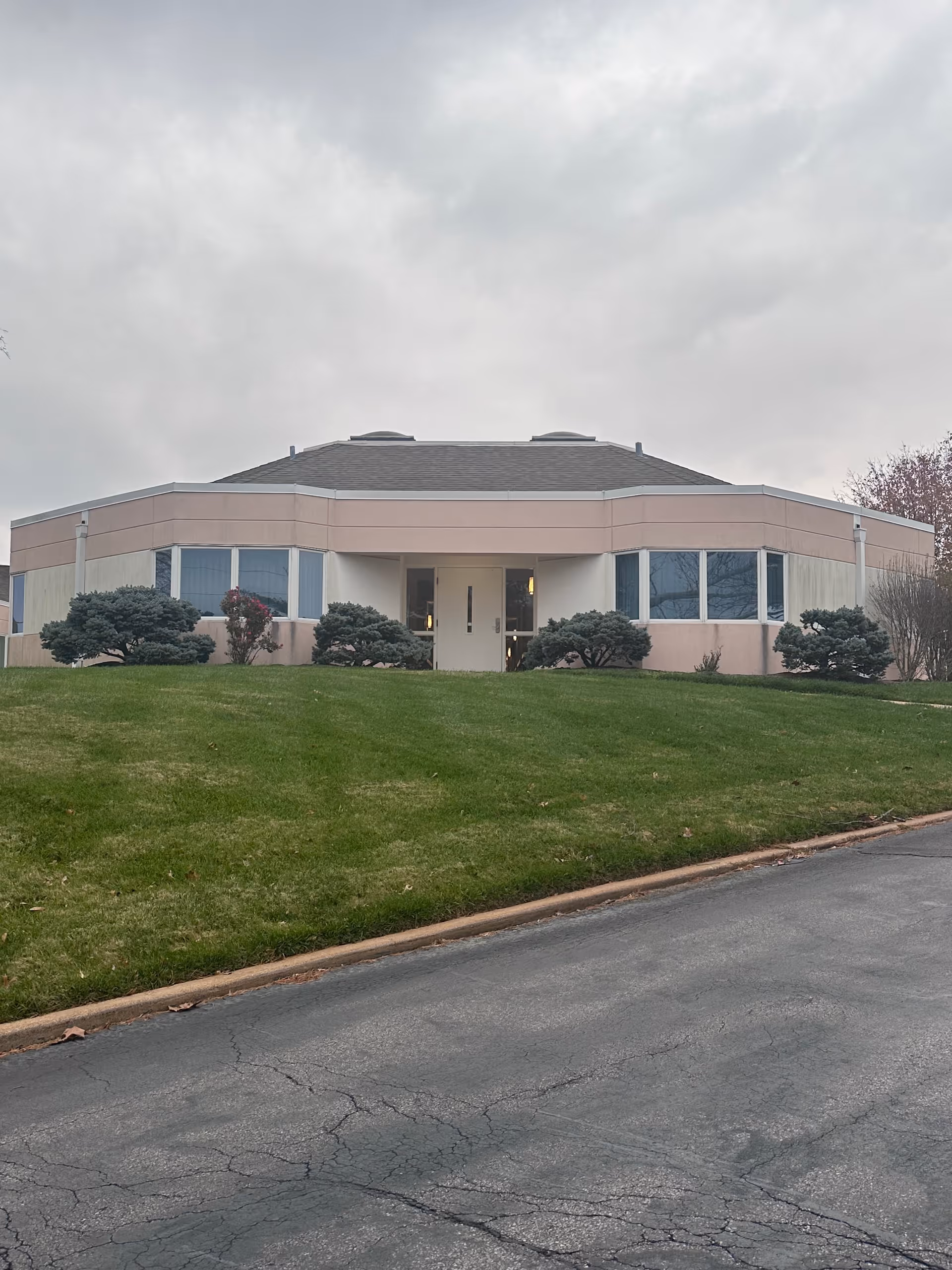 Exterior view of a single-story building with a sloped roof, beige and light pink walls, and large windows. The building is surrounded by neatly trimmed bushes and a well-maintained green lawn. The sky is overcast and the paved driveway in front shows some cracks.