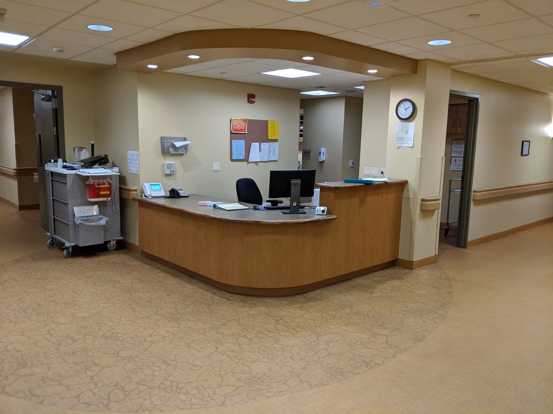 Reception desk area inside Claremont Nursing & Rehabilitation Center with a computer, phone, and office supplies on the desk. There is a clock on the wall and a medical cart with supplies nearby. The hallway extends to the left and right of the desk.