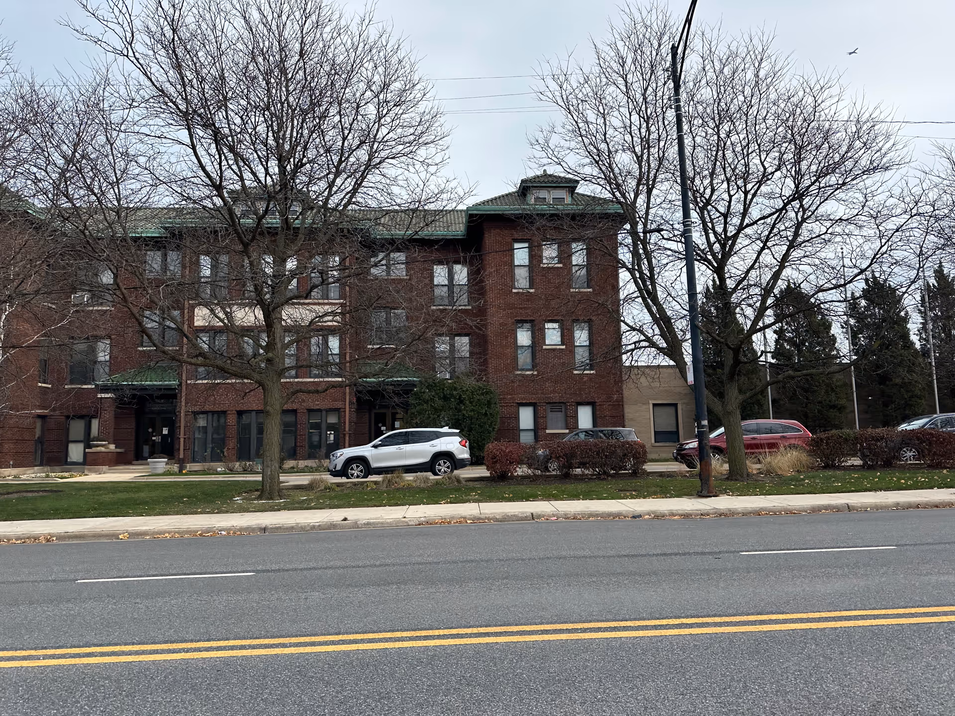 Exterior view of a three-story brick building with multiple windows and a green roof. There are leafless trees and parked cars in front of the building, with a road and double yellow lines in the foreground under a cloudy sky.