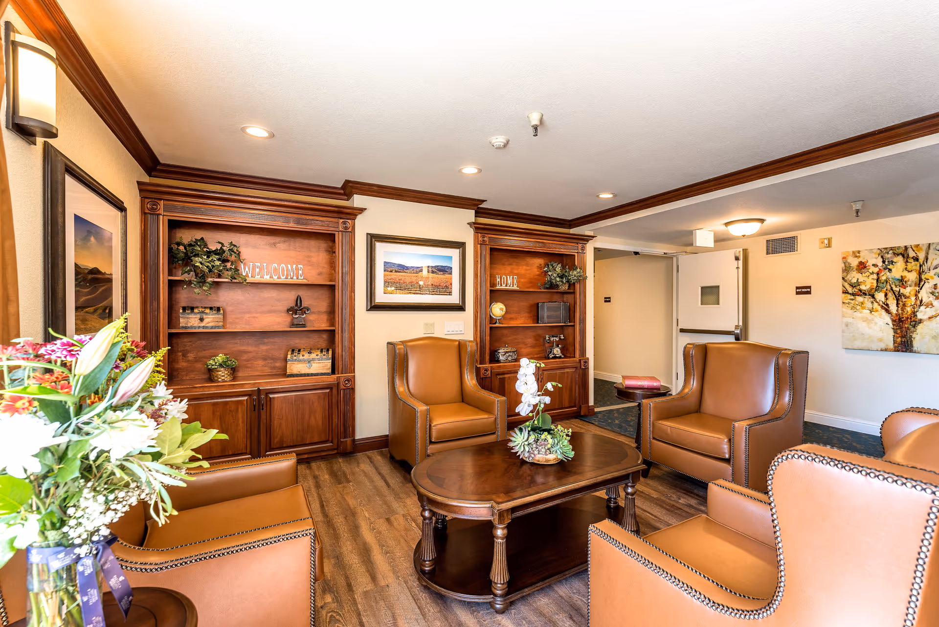 A cozy senior living facility lounge area with four brown leather armchairs arranged around a wooden coffee table with a floral centerpiece. The room features wooden built-in shelves with decorative items and signs that say 'WELCOME' and 'HOME'. There are framed pictures on the walls, a bouquet of flowers on a side table, and warm lighting from wall sconces and ceiling lights.
