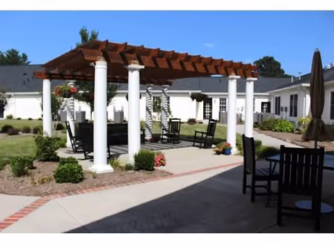Outdoor patio area with a wooden pergola supported by white columns, surrounded by chairs and small plants. The area is part of a senior living facility with white buildings and a well-maintained lawn in the background.