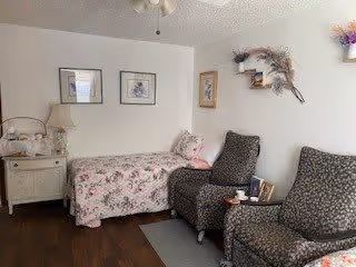 A cozy assisted living room with a single bed covered in floral bedding, two patterned armchairs with a small table between them holding a cup and a book, a nightstand with a lamp and decorative items, framed artwork on the walls, and a decorative wall hanging with flowers.