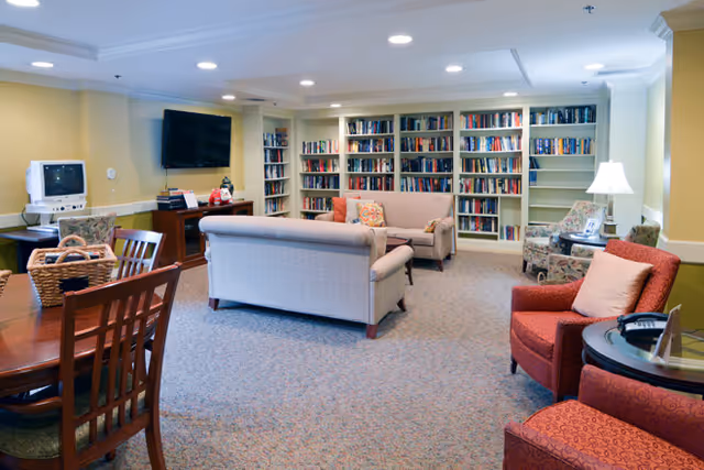 Bright common room with sofas, armchairs, bookshelves, a TV, and a dining table.