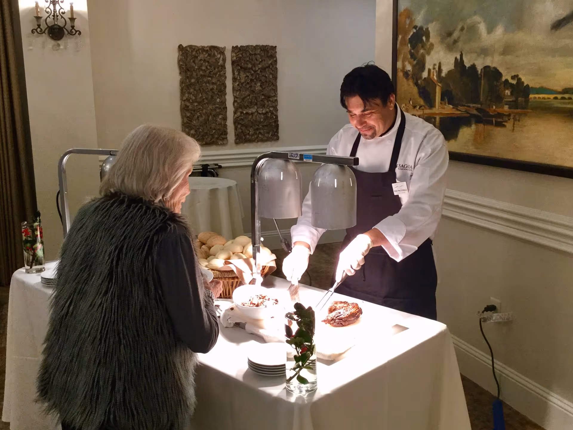 An elderly woman in a dark fur vest is being served food by a chef wearing a white coat and black apron at a buffet table with heat lamps. The setting is an indoor dining area with decorative wall art and a large painting in the background.
