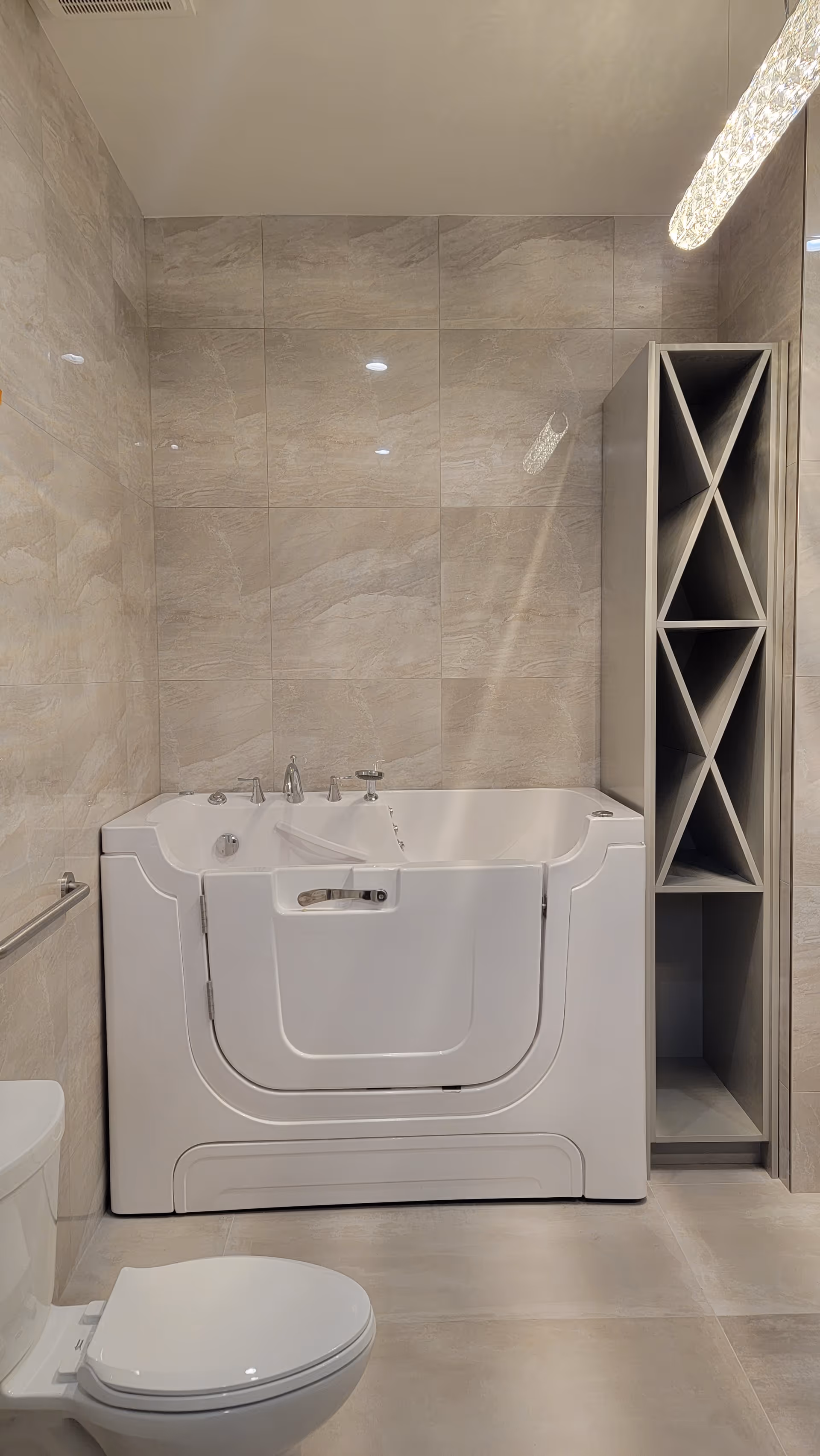 A modern bathroom featuring a walk-in bathtub with a door for easy access, beige tiled walls and floor, a toilet, and a tall shelving unit with multiple compartments.