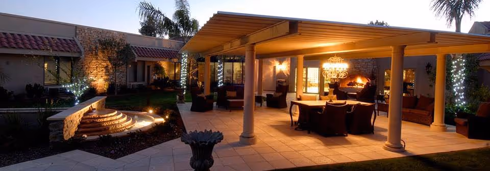 Dusk view of a landscaped courtyard with a covered patio featuring a dining table and lounge seating, a lit fountain, and trees wrapped in lights in front of a single-story building.