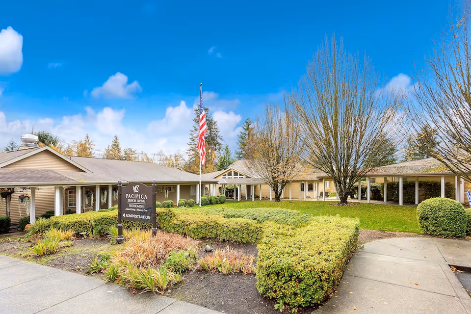 Exterior view of The Cottages of Snohomish senior living facility showing a single-story building with beige siding, a well-maintained garden with trimmed bushes and trees, a flagpole with an American flag, and a sign for Pacifica Senior Living Snohomish Administration.