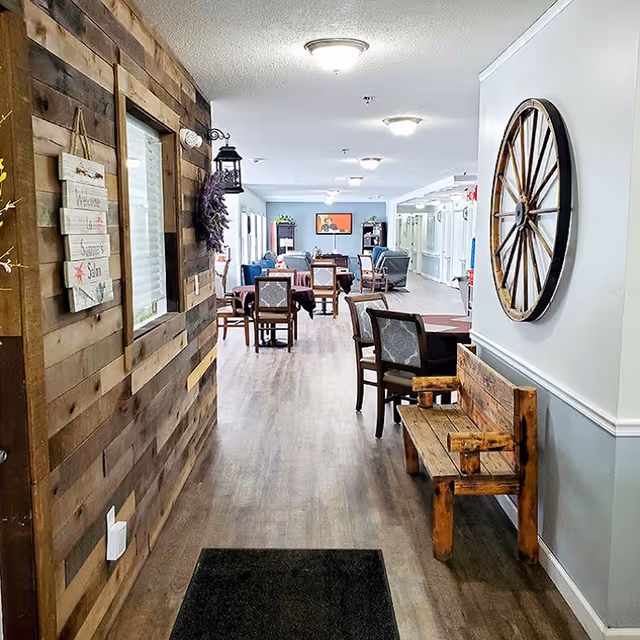 A hallway in a senior living facility with wood-paneled walls on the left and light gray walls on the right. A rustic wooden bench is placed against the right wall beneath a large decorative wagon wheel. Several tables and chairs are arranged further down the hallway, leading to a common sitting area with armchairs and a TV mounted on the far wall. The floor is wood laminate, and ceiling lights illuminate the space.