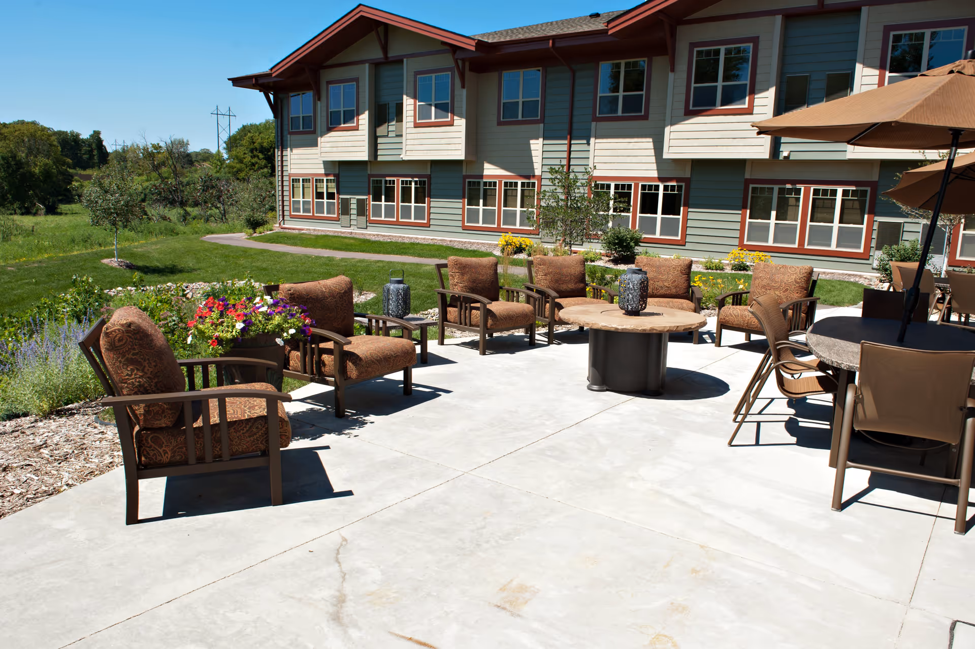 Outdoor patio area with cushioned chairs arranged around a circular table and additional seating with umbrellas. The patio is adjacent to a two-story building with multiple windows and surrounded by greenery and flowers under a clear blue sky.
