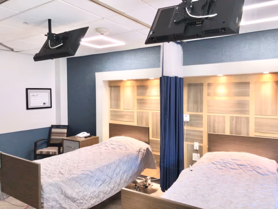 A room with two hospital-style beds separated by a blue privacy curtain. Each bed has a light gray quilted blanket. Above the beds, there are mounted flat-screen televisions on the ceiling. The wall behind the beds features a decorative wood panel design with built-in lighting. A chair and a small table with a telephone are visible in the corner of the room.