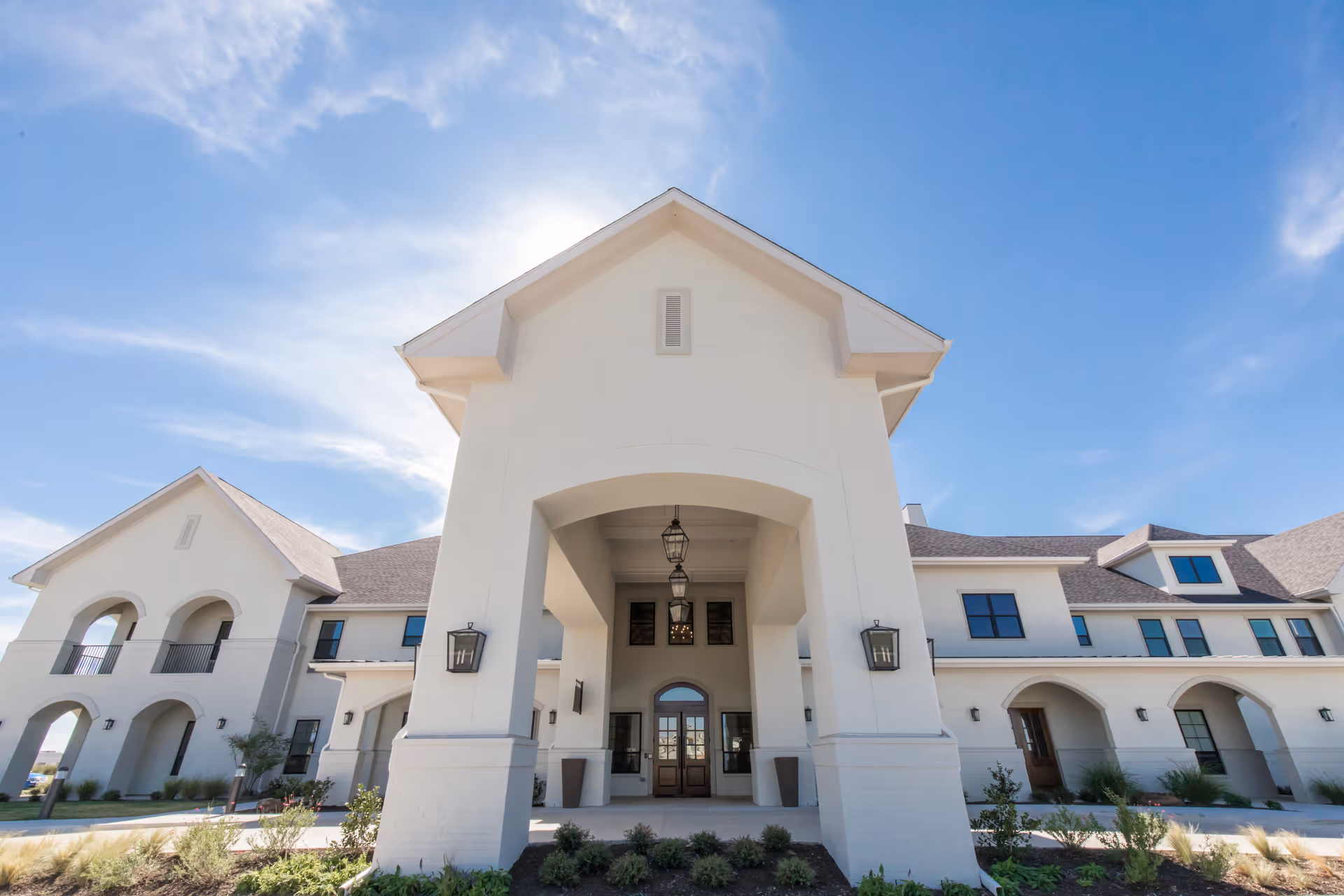 Exterior front view of a large, modern senior living facility building with white walls, multiple windows, and an arched entrance under a clear blue sky.