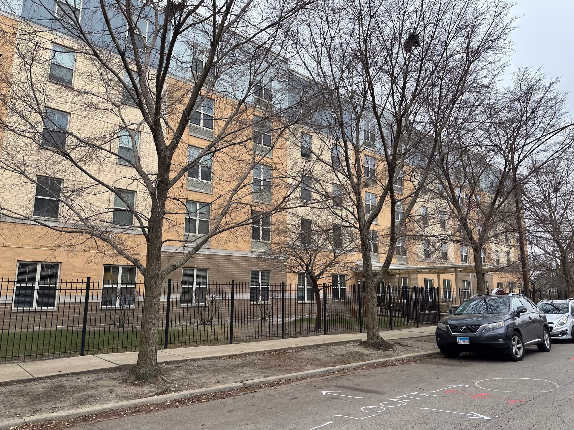 Front exterior of a multi-story brick residential building with leafless trees and parked cars along the street.
