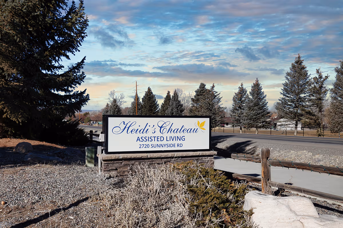Outdoor sign for "Heidi's Chateau Assisted Living" by a roadside with trees and landscaping under a partly cloudy sky.