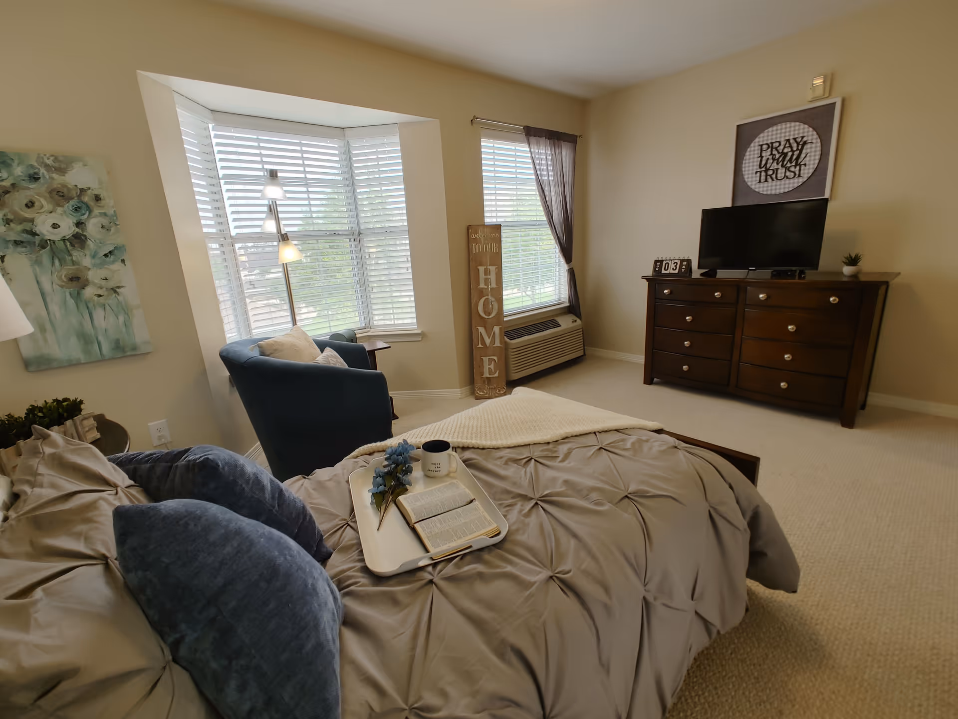 A cozy bedroom with a bed covered in gray bedding and blue pillows. On the bed is a tray holding a book, a mug, and a small bouquet of flowers. A blue armchair sits near a bay window with white blinds and a floor lamp. A wooden dresser with a TV on top is against the far wall, with a framed sign above it that reads 'PRAY, WAIT, TRUST'. A decorative wooden sign leaning against the wall near the window says 'HOME'. The room has beige walls and carpeted floor.