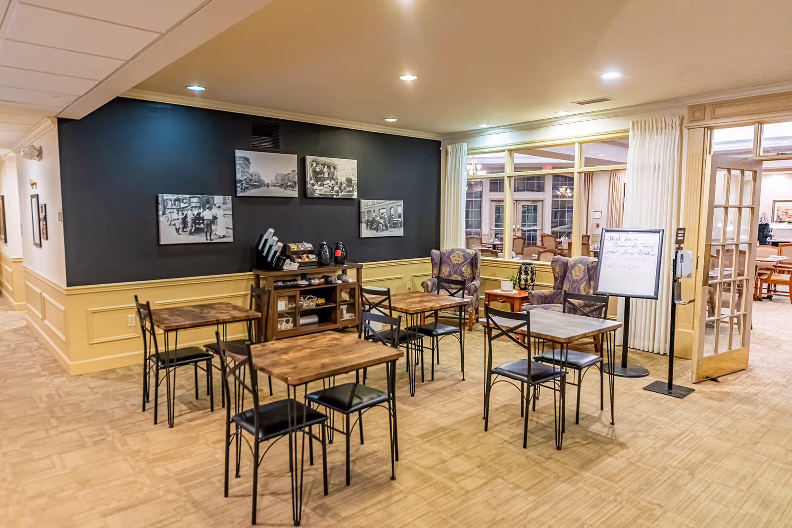 A cozy common area with several small wooden tables and black metal chairs arranged on a carpeted floor. There are two upholstered armchairs with floral patterns near a windowed wall that looks into another room with more seating. A black accent wall displays several black and white photographs above a wooden shelving unit holding coffee supplies. A whiteboard on a stand is placed near the entrance to the adjacent room.