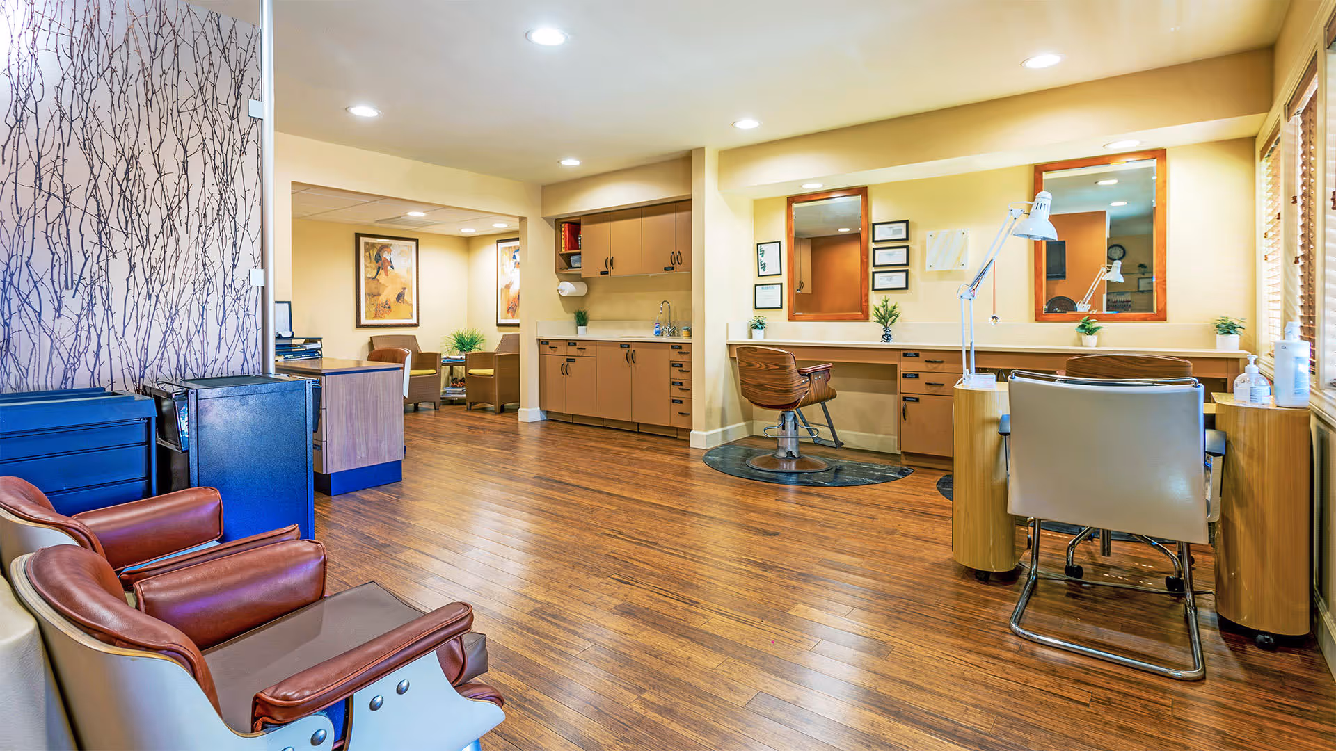 Interior view of a salon area in a senior living facility with wooden flooring, salon chairs, mirrors, and workstations. There are also some seating chairs and a small desk in the background, with soft yellow walls and decorative plants.