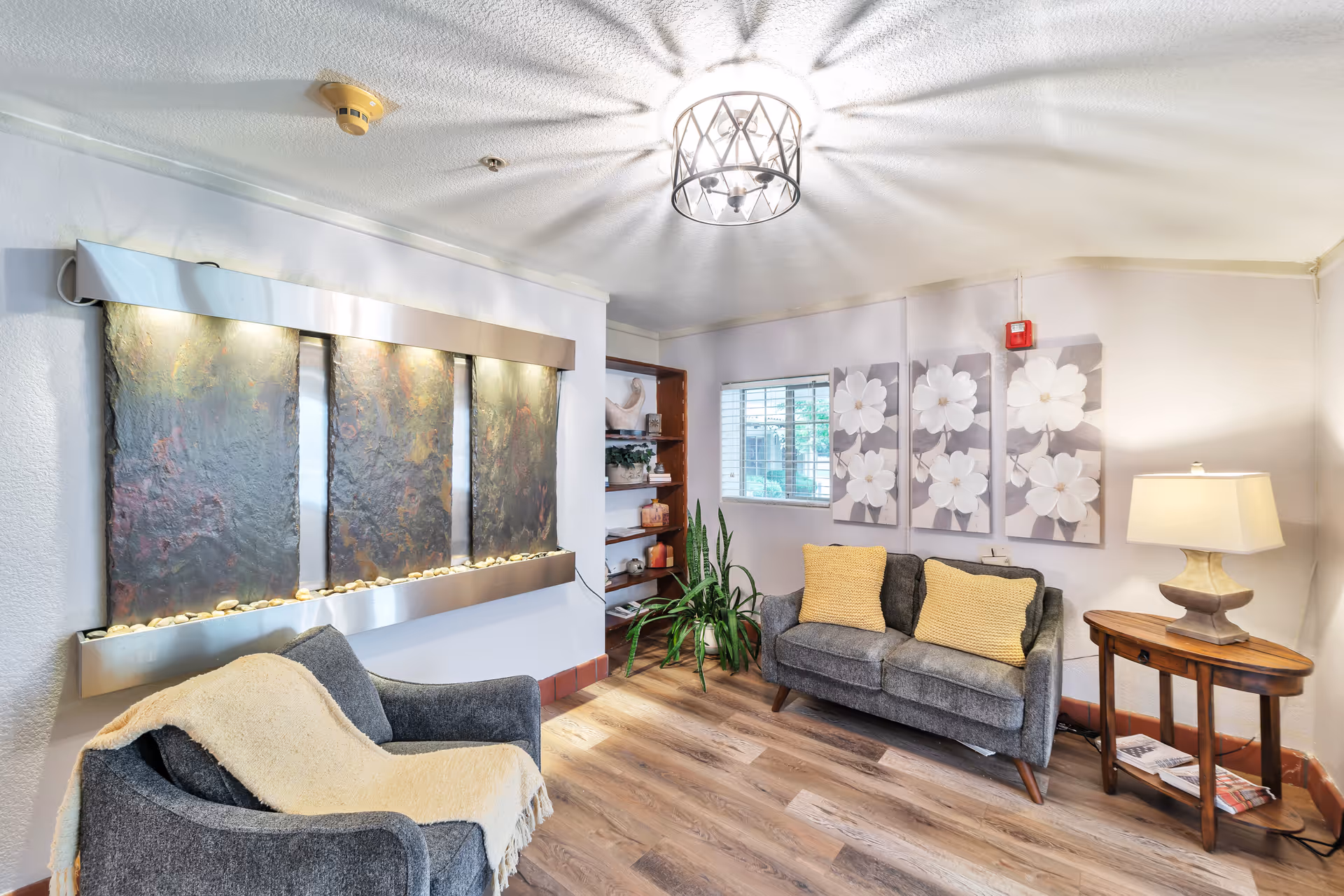 A cozy senior living facility common area with two gray upholstered chairs, one with a beige throw blanket, and a matching loveseat with two yellow cushions. A wooden side table with a lamp and magazines is beside the loveseat. On the wall above the loveseat are three large floral paintings. A decorative water feature with three vertical panels and small stones is mounted on the opposite wall. The room has wood flooring, a ceiling light fixture, and a window letting in natural light.