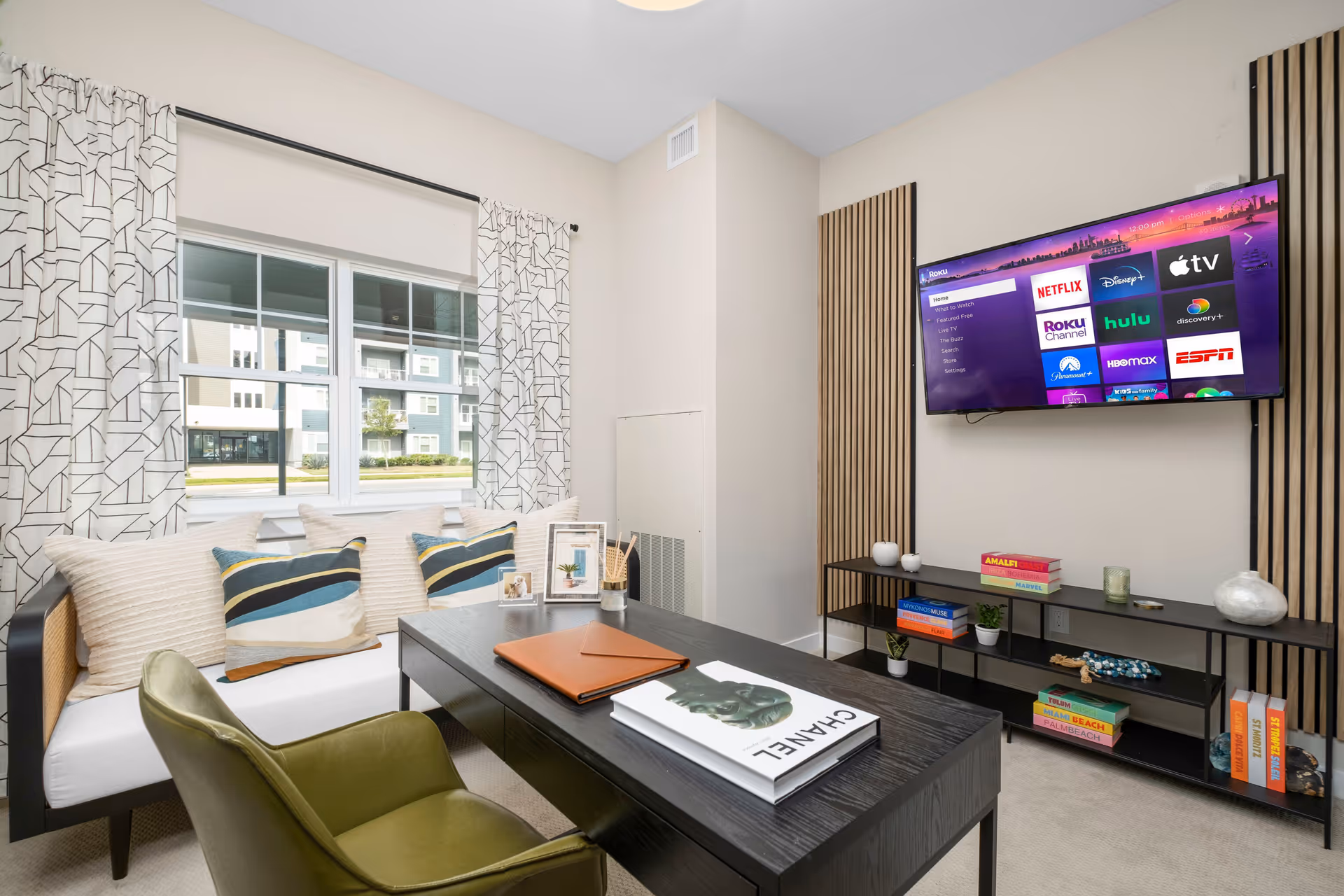 A modern living room with a black coffee table holding books and a leather folder, an olive green armchair, and a white cushioned bench with decorative pillows. A large flat-screen TV is mounted on the wall displaying streaming service options. The room has light-colored walls, a window with patterned curtains, and a black shelving unit beneath the TV with decorative items and books.