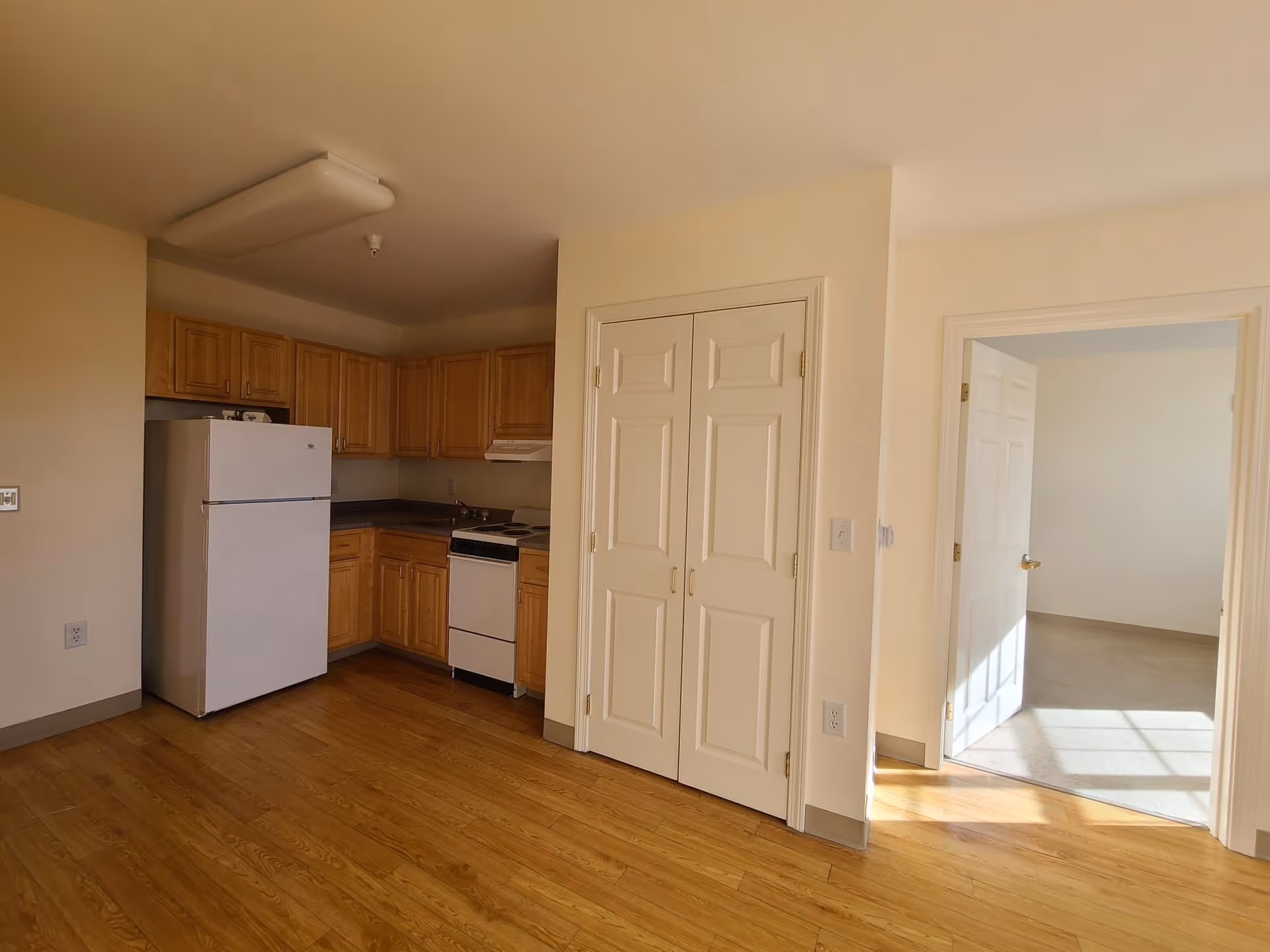 A small kitchen area with wooden cabinets, a white refrigerator, a white stove, and a double-door pantry. The kitchen has wood flooring and beige walls. To the right, there is an open doorway leading to a carpeted room with sunlight streaming in.