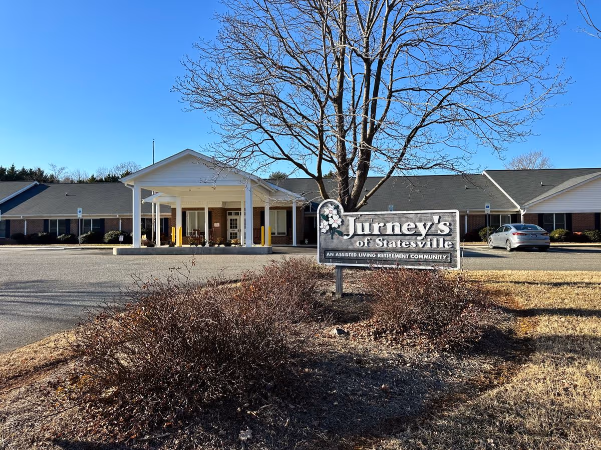 Front exterior of Jurney's of Statesville assisted living facility with an entrance portico, parking lot, and a sign in the foreground.