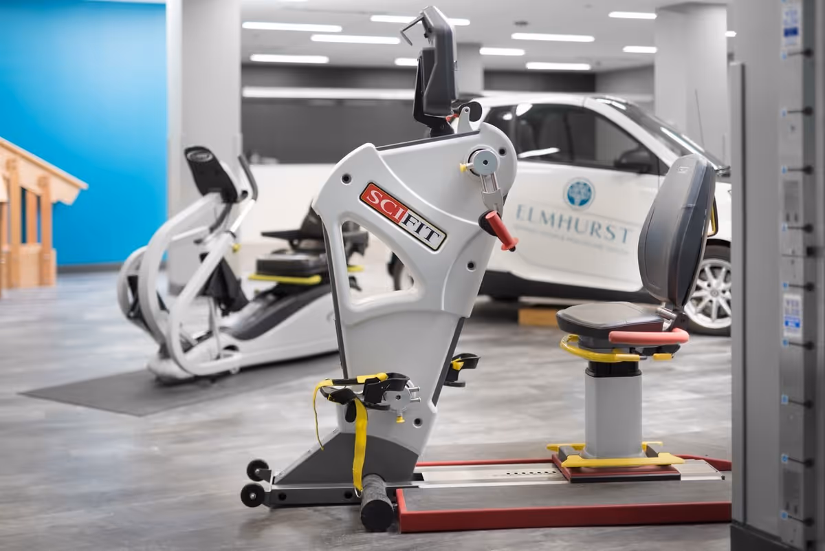 Indoor rehabilitation gym area with exercise equipment including a SciFit machine and a recumbent bike. In the background, there is a white car with the Elmhurst Rehabilitation & Healthcare Center logo on the door. The room has a blue accent wall and bright overhead lighting.