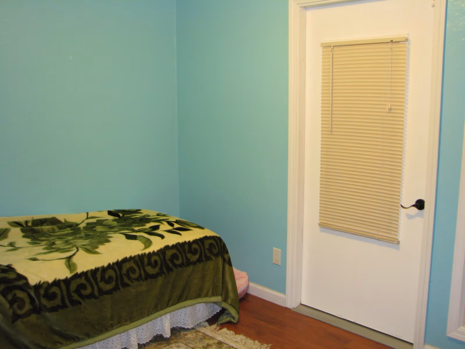 A small bedroom with light blue walls, a bed covered with a green floral blanket, a white door with closed beige blinds, and wooden flooring.