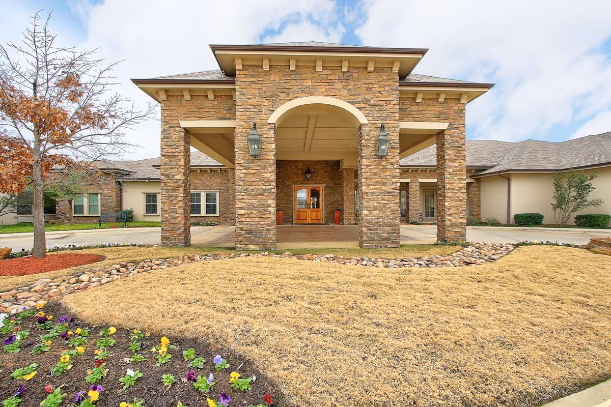 Front exterior view of The Oaks At Flower Mound facility featuring a stone entrance with large pillars, a covered entryway, and landscaped grounds with grass, flowers, and small trees under a partly cloudy sky.