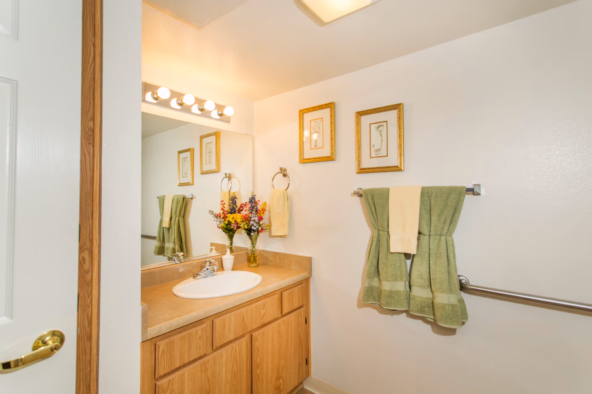 A bathroom with a wooden vanity cabinet, a countertop with a white sink, and a large mirror above it. There are four round light bulbs above the mirror. Two framed pictures hang on the wall above a towel rack holding two green towels with a smaller beige towel in the middle. A vase with colorful flowers and a soap dispenser are on the countertop. A metal grab bar is mounted on the wall beside the towel rack.