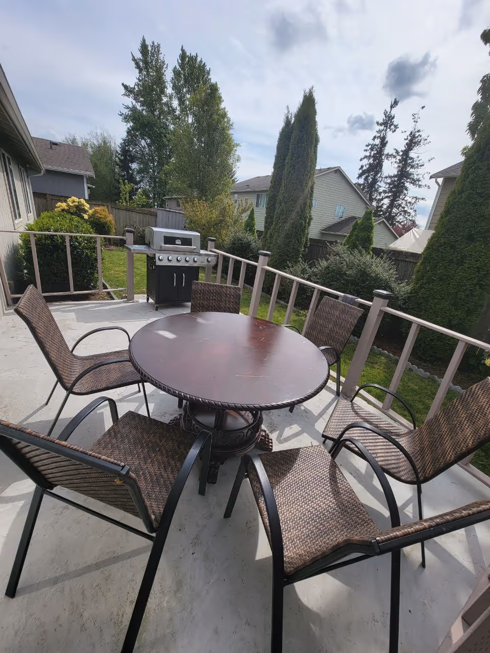 Outdoor patio area with a round wooden table surrounded by six wicker chairs. In the background, there is a gas grill and a fenced yard with green bushes, tall trees, and neighboring houses under a partly cloudy sky.